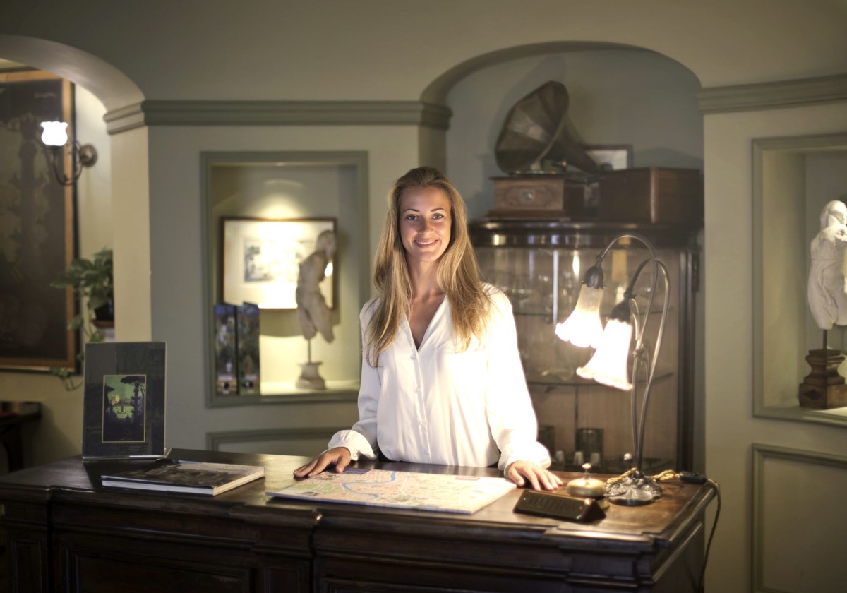A smiling receptionist at the front desk of a boutique hotel, with greetings in English, Spanish and French floating above her.